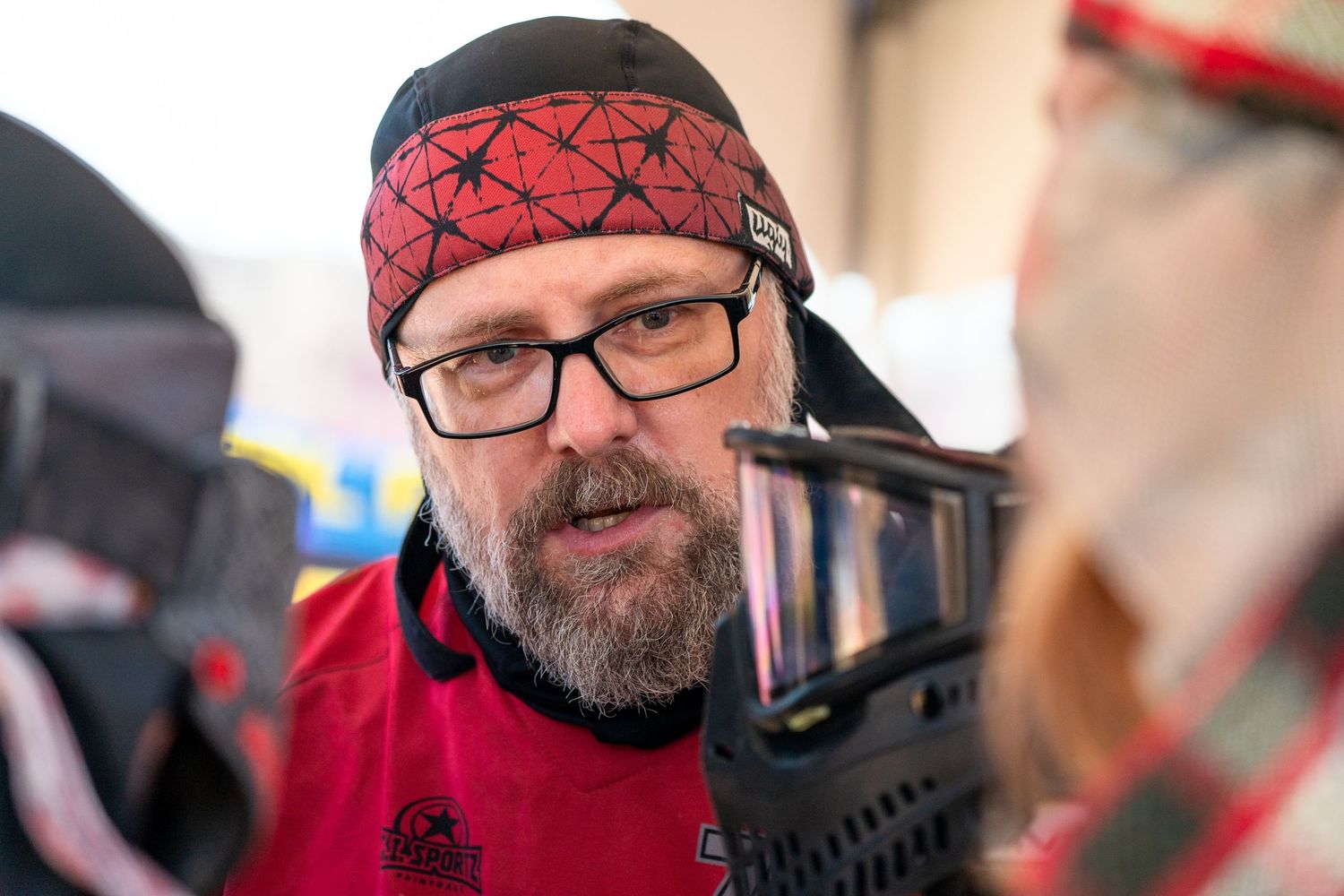 Man with glasses and red bandana wearing red jersey near paintball mask outdoors.
