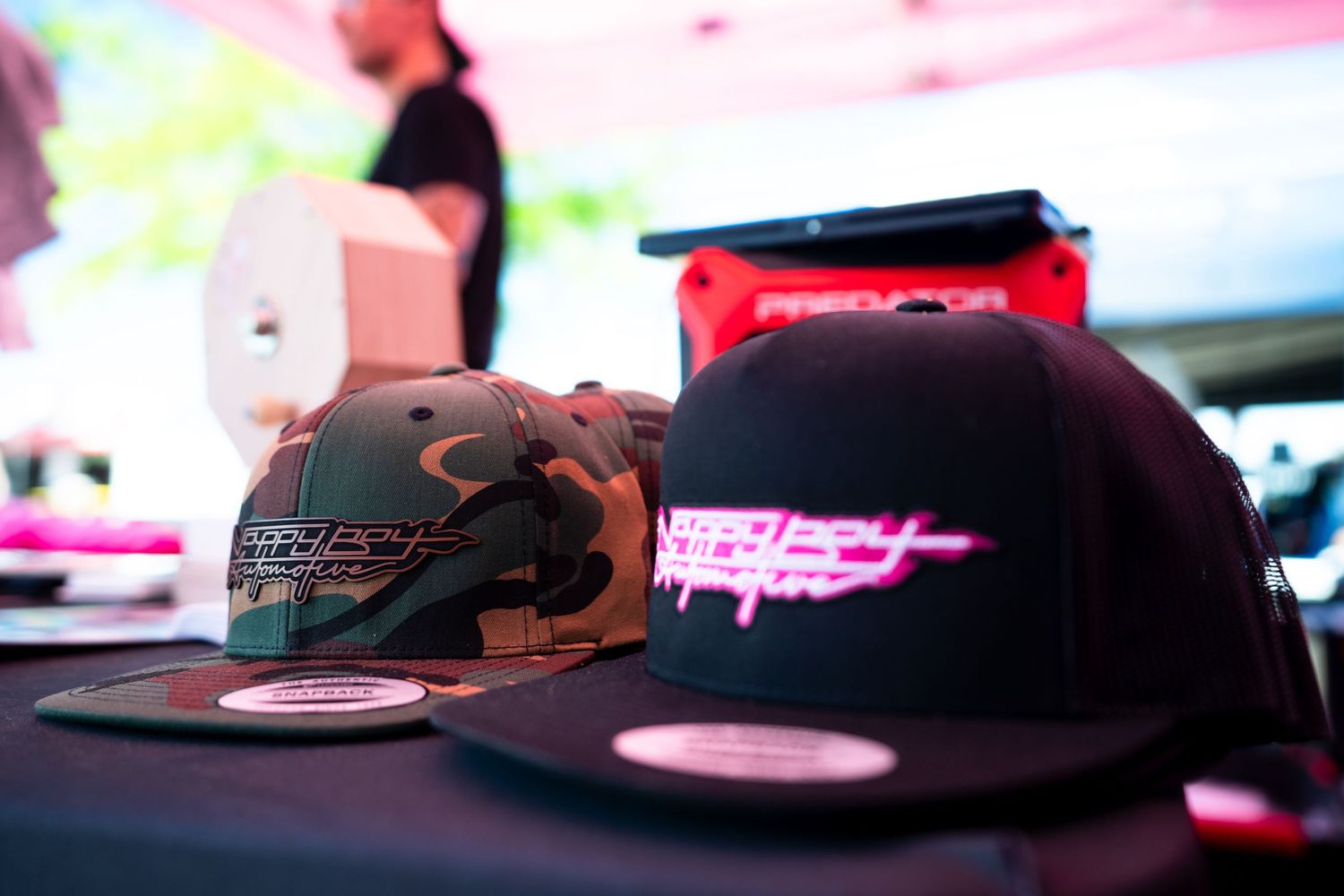 Two snapback hats with colorful logos displayed on a table at a motorsport merchandise booth.