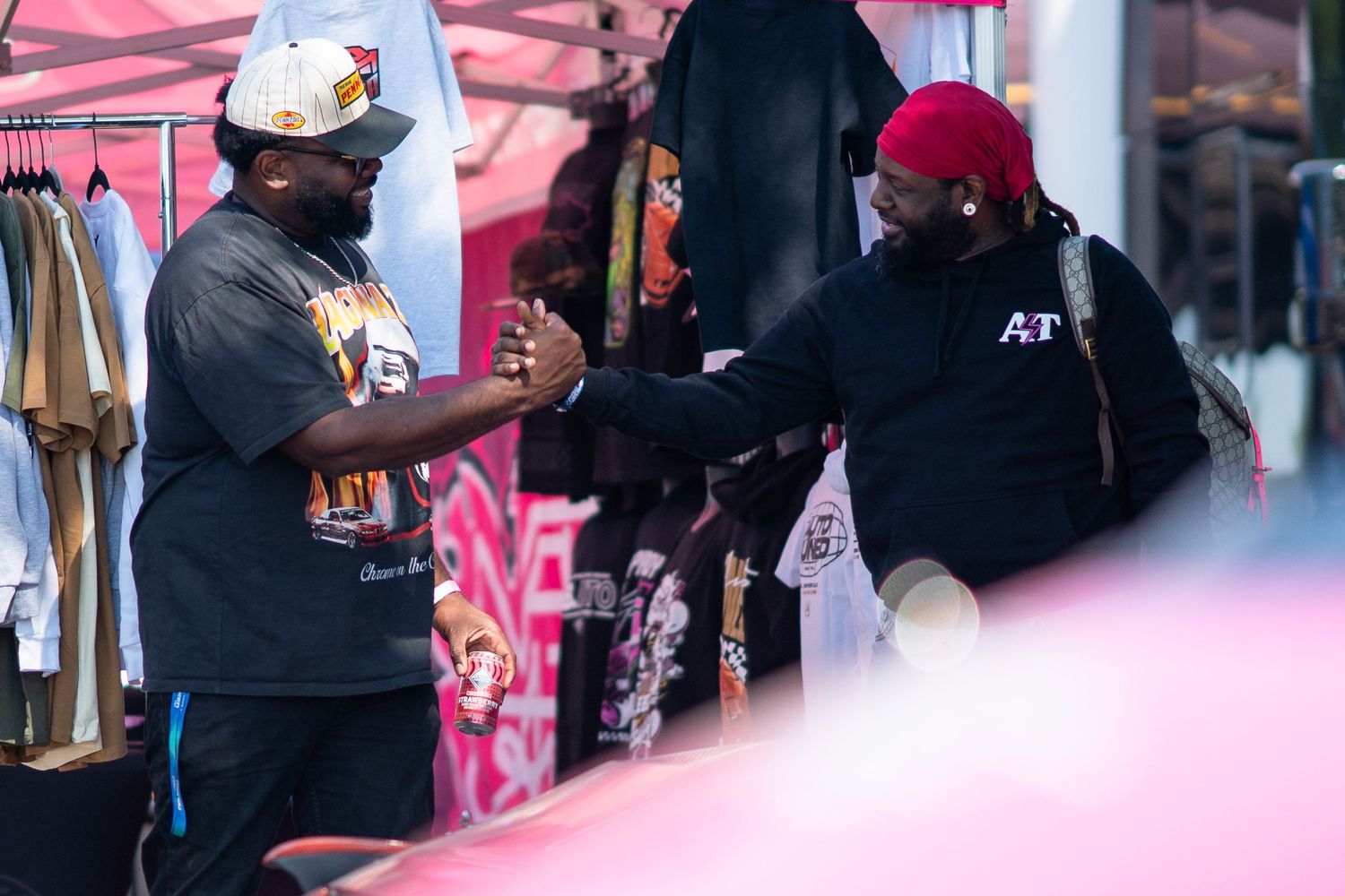 Two men fist bumping at an outdoor market vendor booth with clothing displayed in the background.