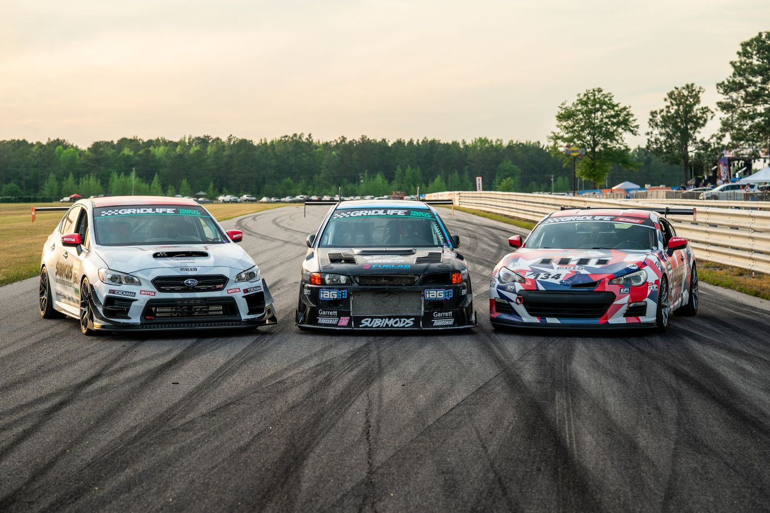Three modified Subaru race cars lined up on a track at dusk, ready for motorsport competition.