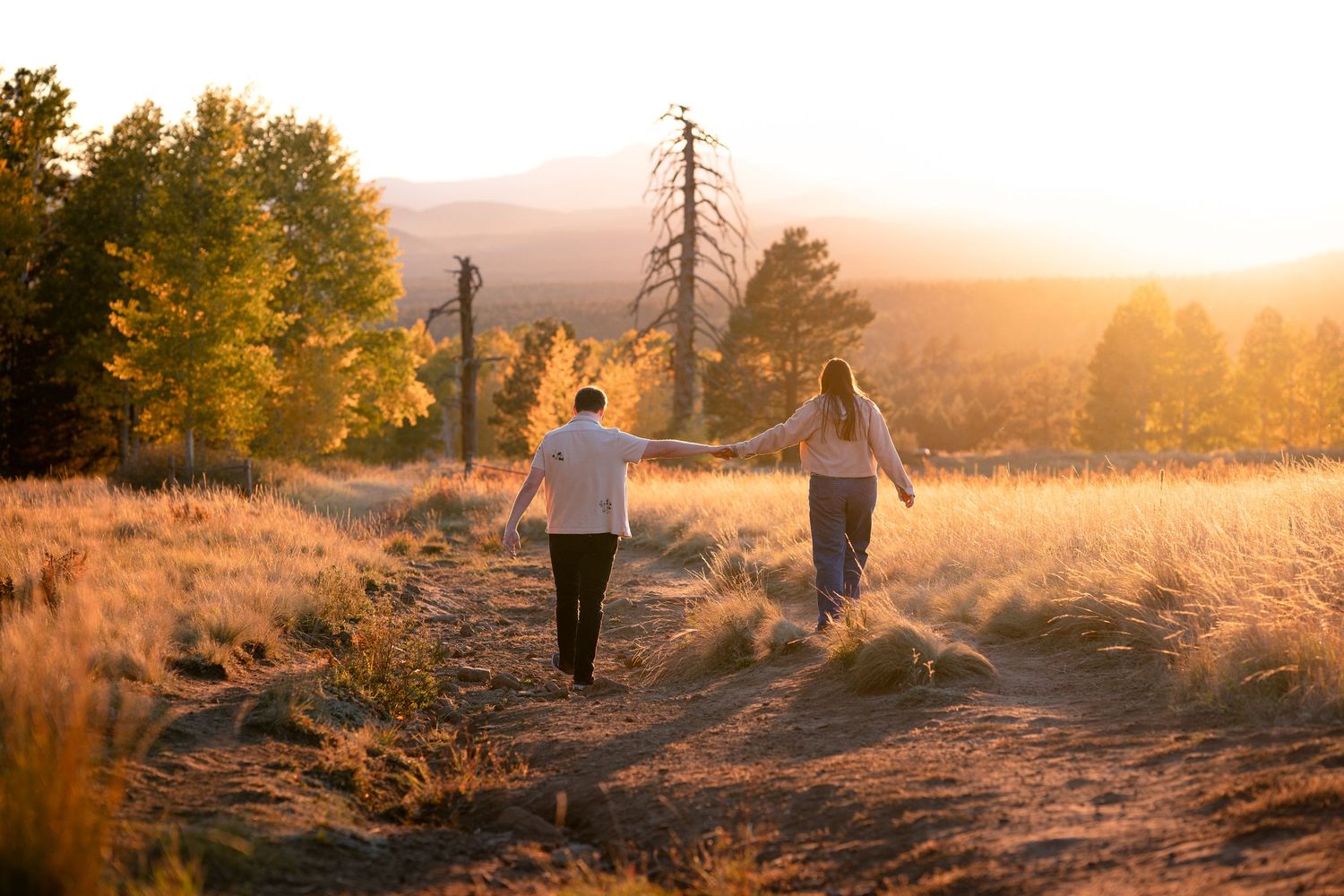 Couple holding hands while walking along a dirt path through golden grass and autumn trees at sunset in a scenic mountain landscape.