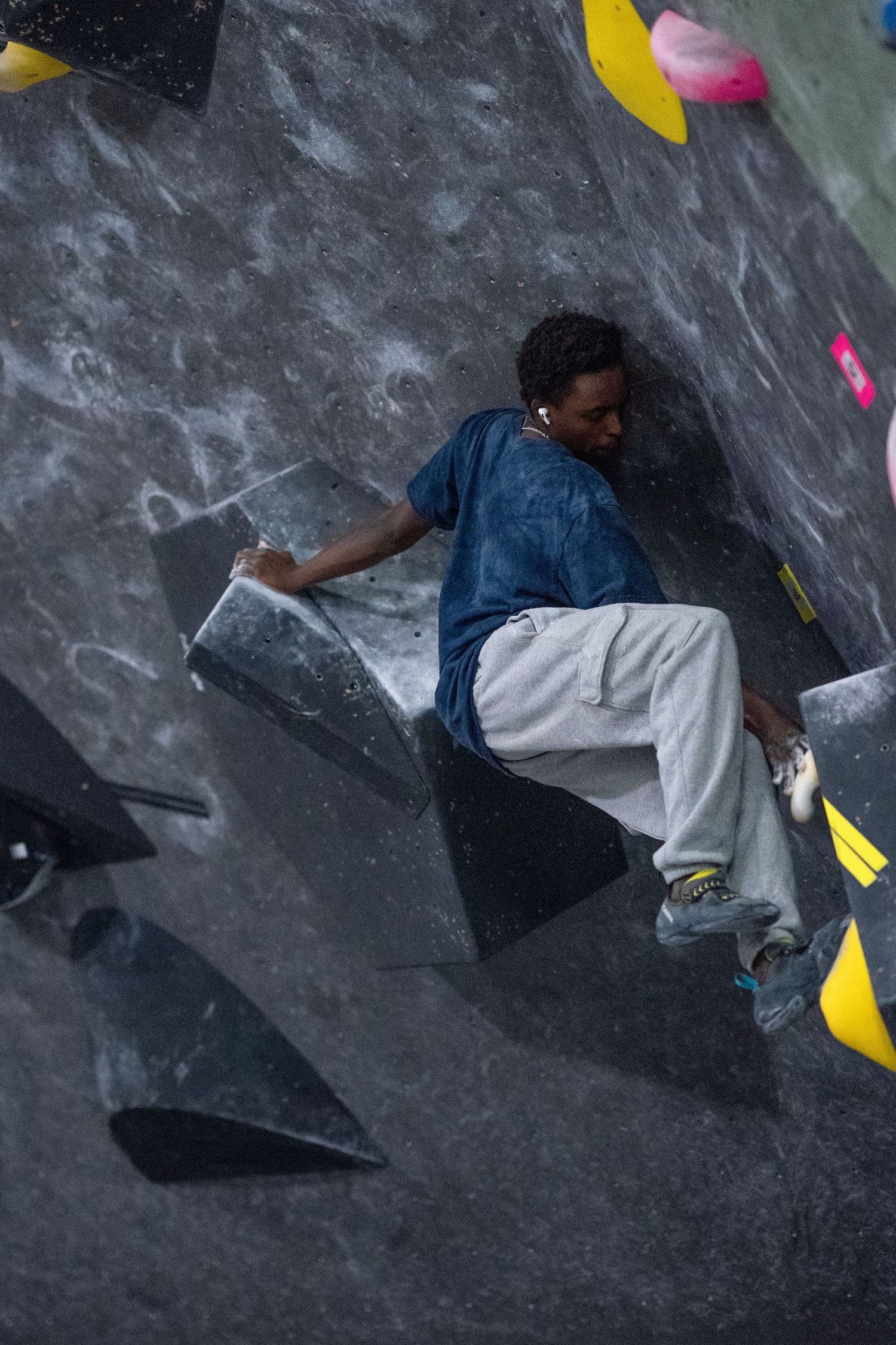 A climber in a blue t-shirt and gray pants balances on a dark angled wall at an indoor bouldering gym.