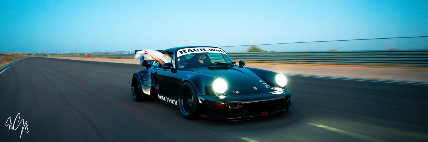 Modified black Porsche 911 in motion with headlights on against blue sky.