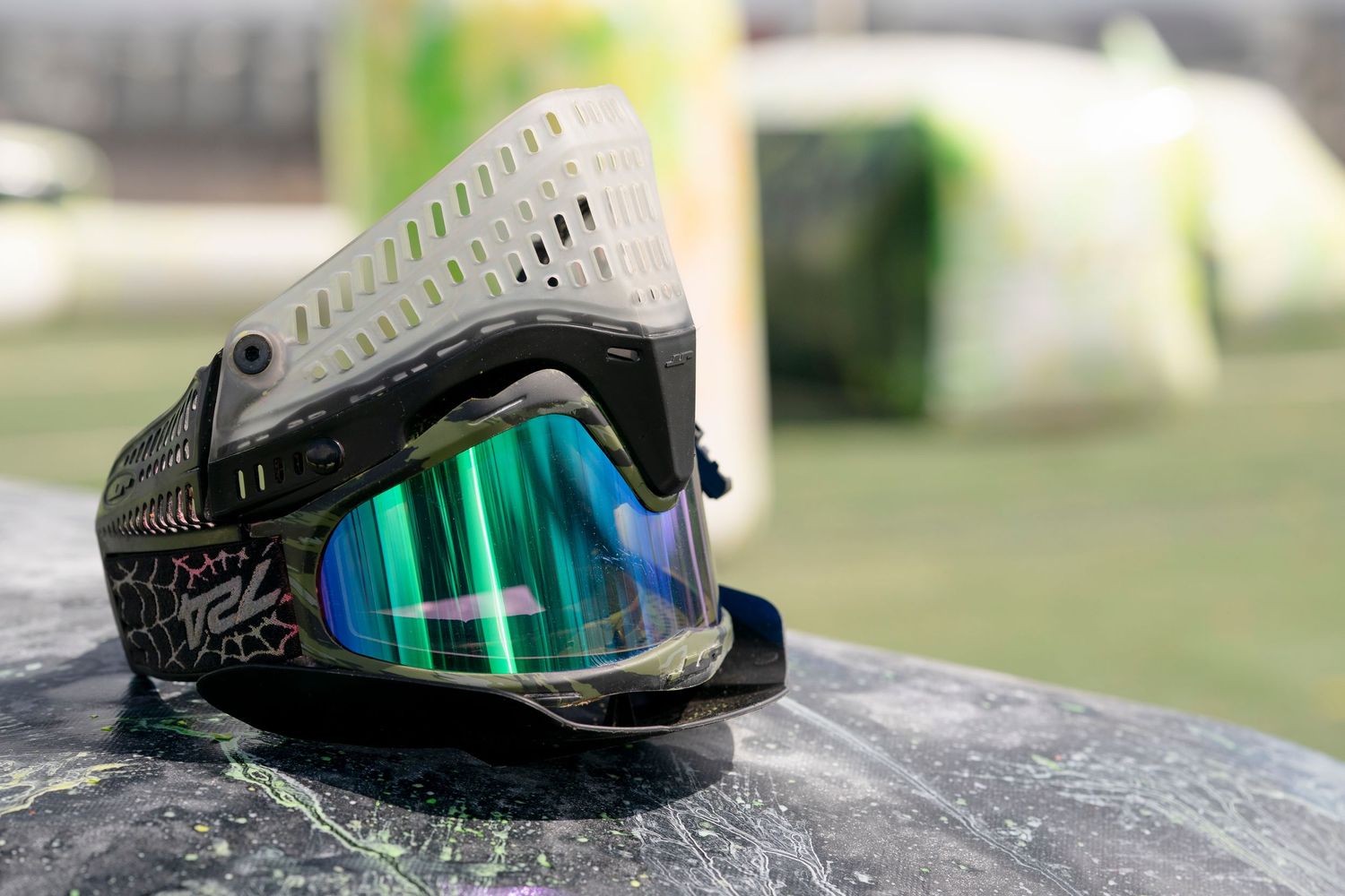 Paintball mask with green mirrored lens sitting on dark surface with bunkers in background.