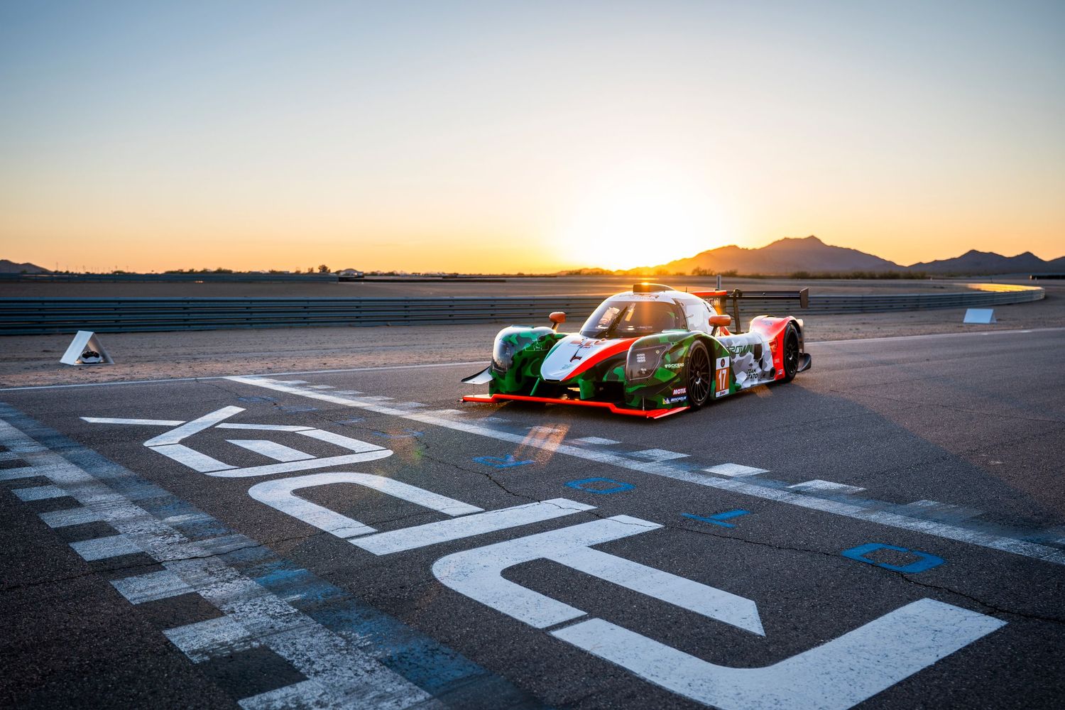 Race car positioned at start/finish line during dramatic sunset with mountains on horizon.