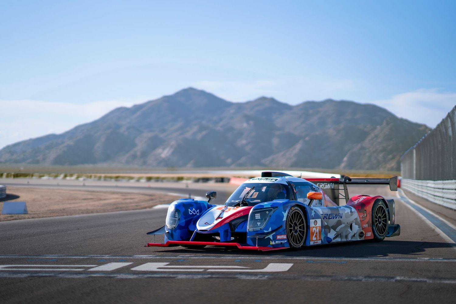 Blue and red race car speeding along a racetrack with mountains visible in the background.