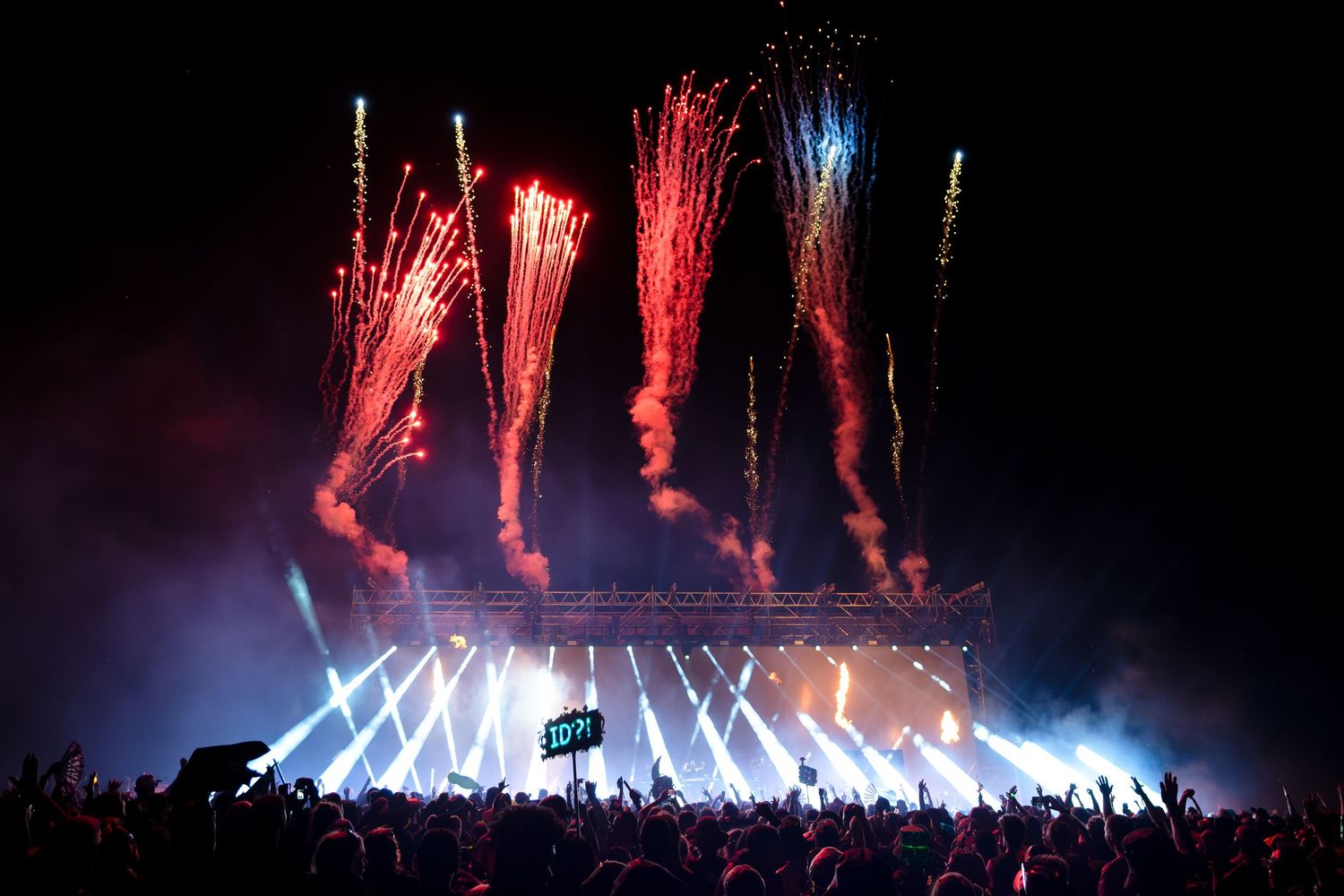 Spectacular fireworks display over a concert stage with bright light beams and silhouetted crowd at night.