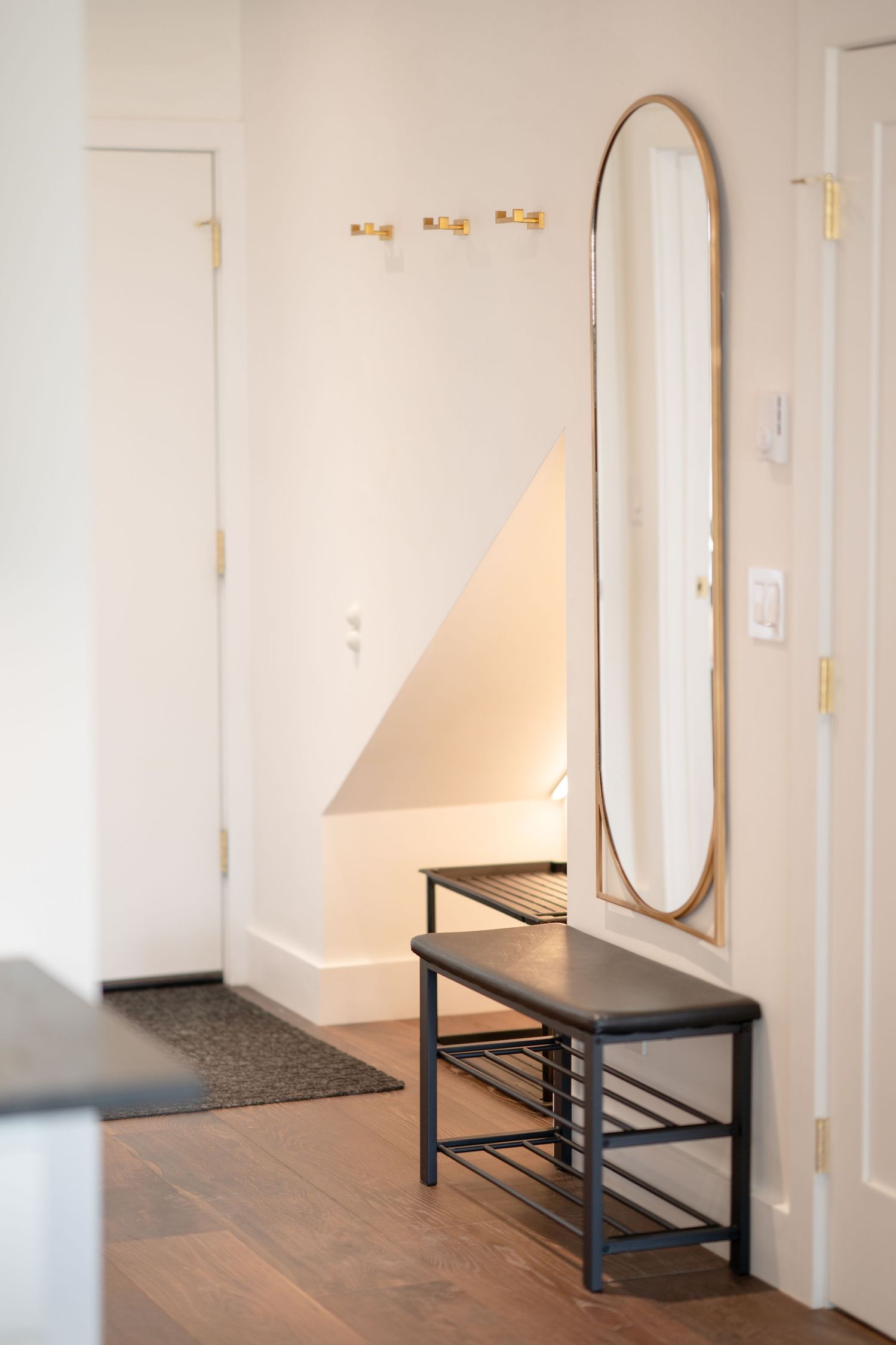 Modern entryway with black shoe bench, tall oval gold-framed mirror, brass wall hooks, and warm hardwood floors.