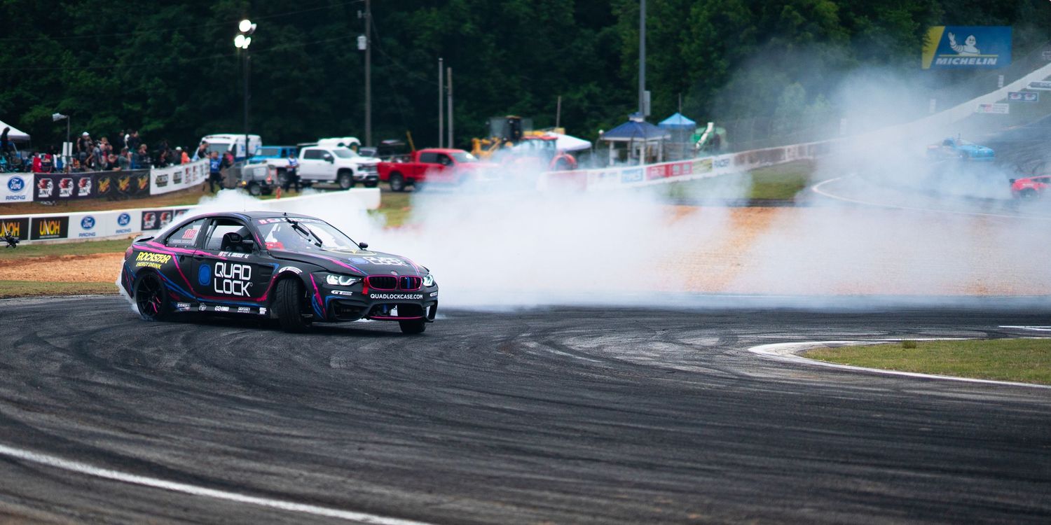 Purple drift car producing massive smoke clouds while drifting on a race track at a motorsport event.