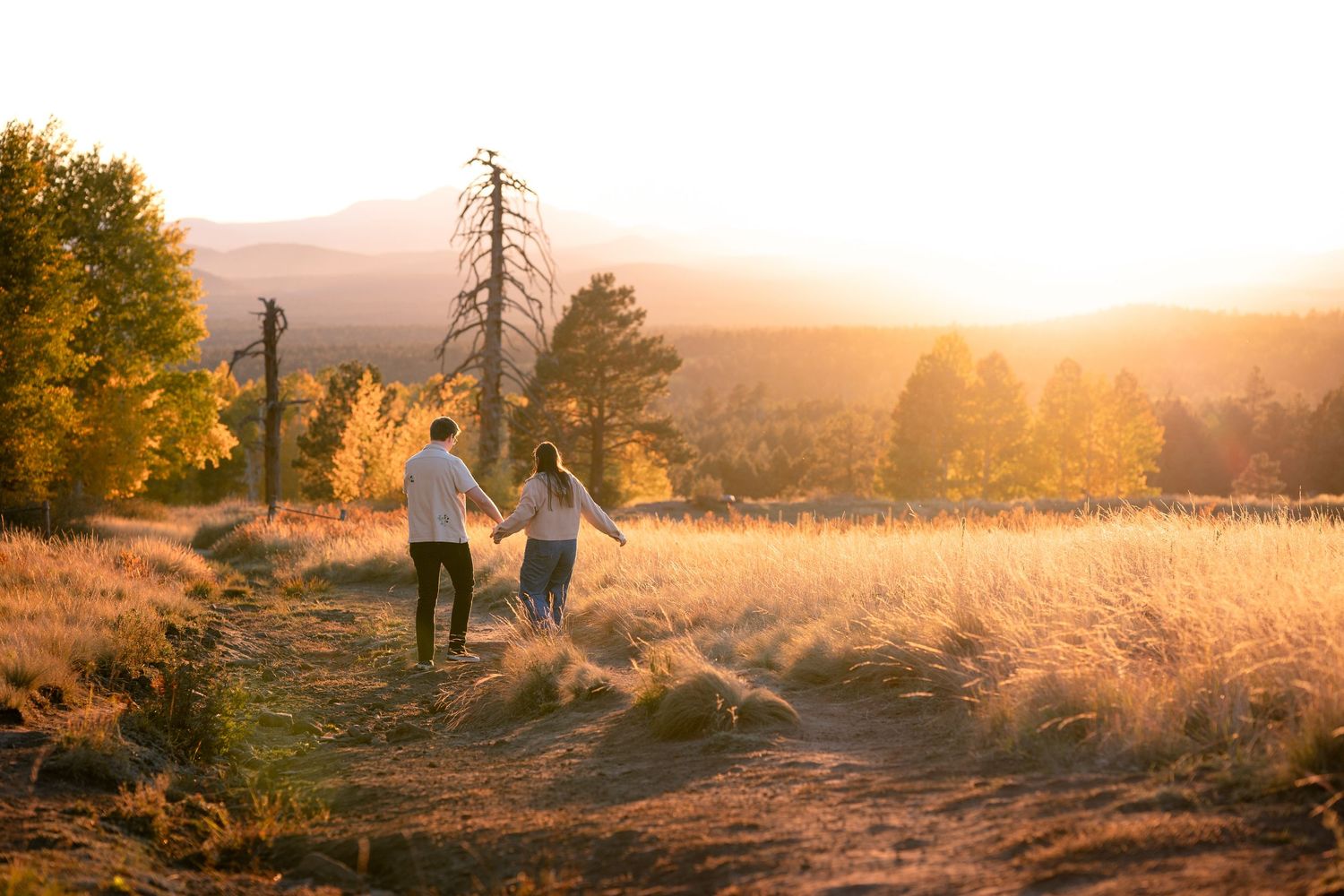 Couple holding hands and walking along a dirt path through golden grass and autumn trees at sunset in the mountains.