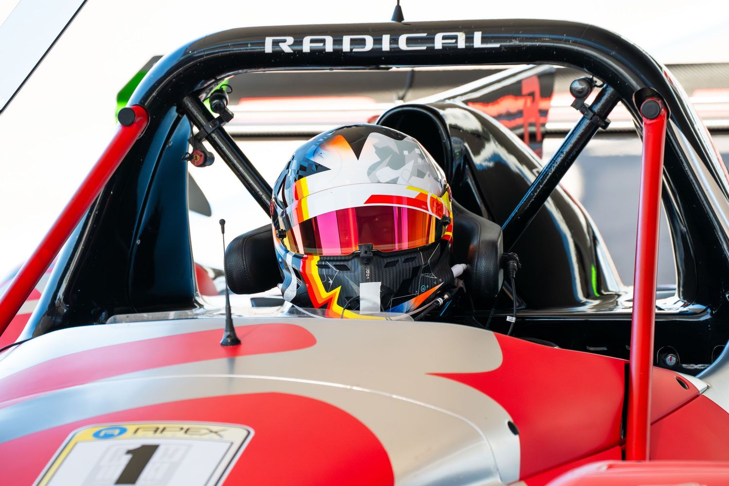 Racing driver in cockpit of red Radical sports car with RADICAL branding visible.