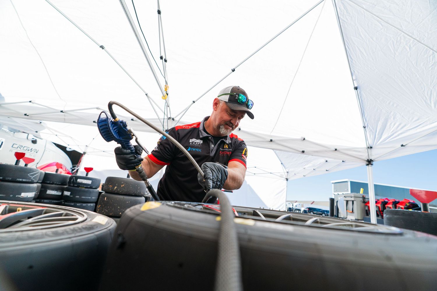 Racing team technicians inspect and work on vehicle components under white canopy tents.