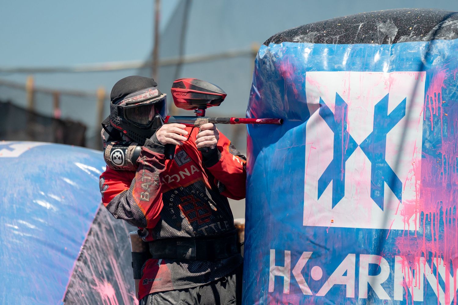 Paintball player in mask aiming marker beside HK Army branded inflatable bunker.