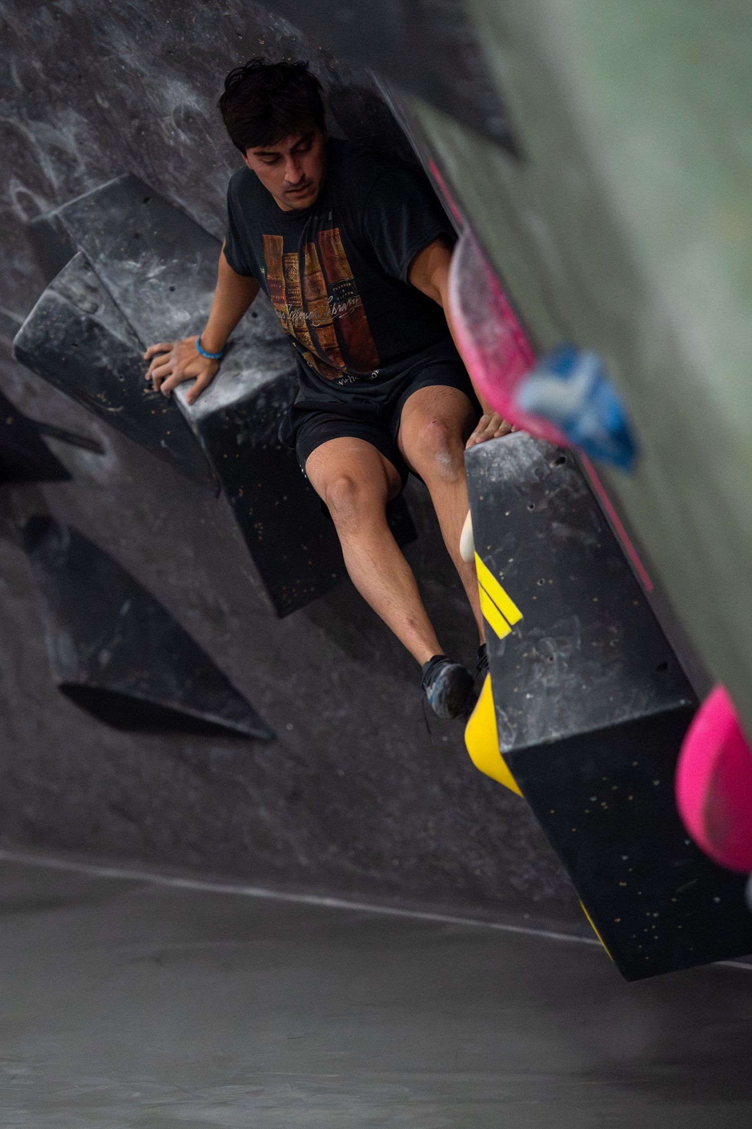 Climber in a dark t-shirt and shorts bouldering on a steep indoor wall with black volumes and colorful holds.