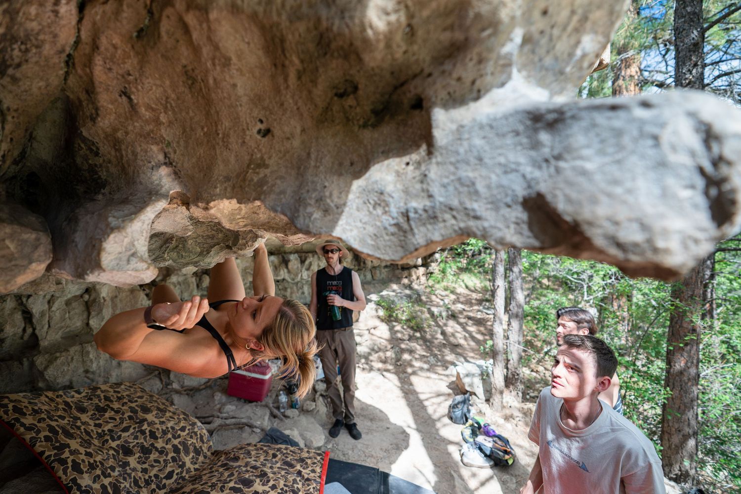 A climber hangs beneath a large rock overhang while a spotter and onlookers watch from a forest clearing with crash pads below.