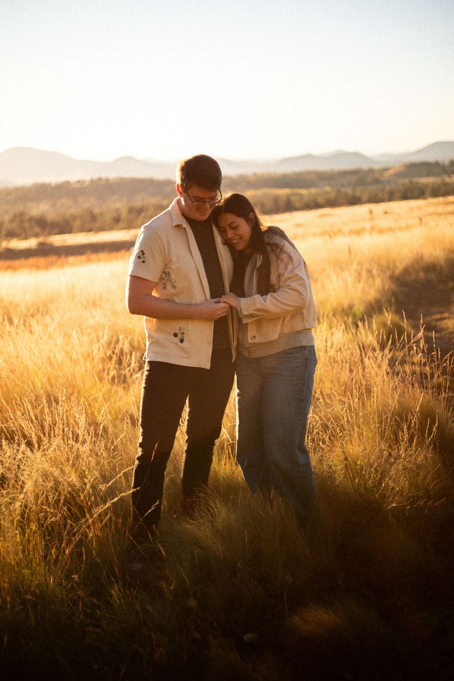 Smiling couple standing close together in tall golden grass at sunset, with distant mountains glowing in the background.