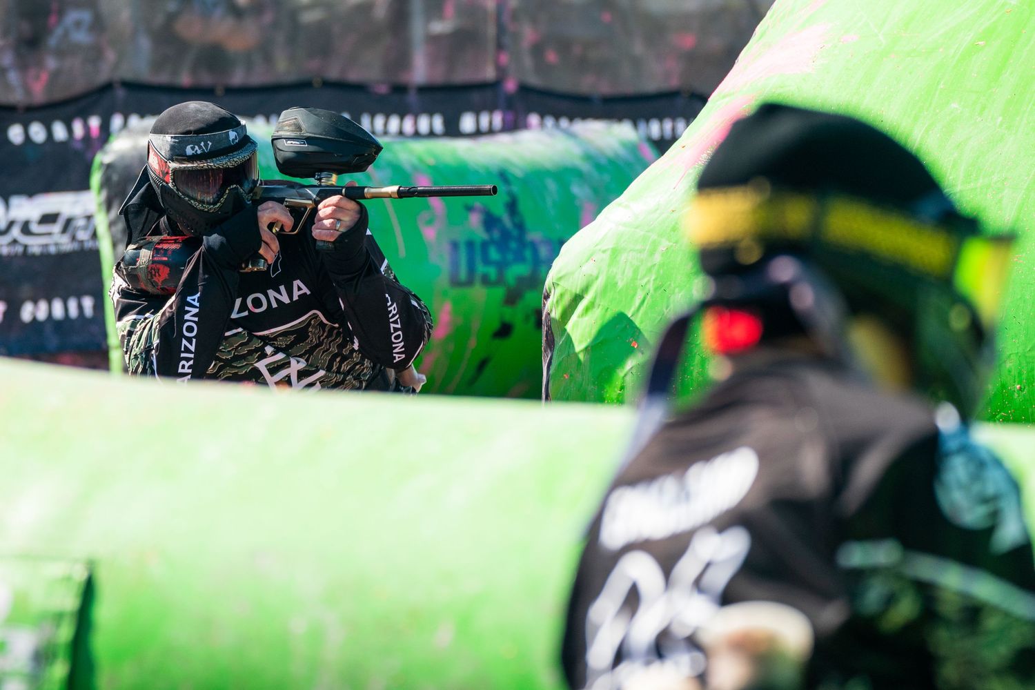 Players in protective gear compete in a paintball tournament behind bright green inflatable bunkers outdoors.