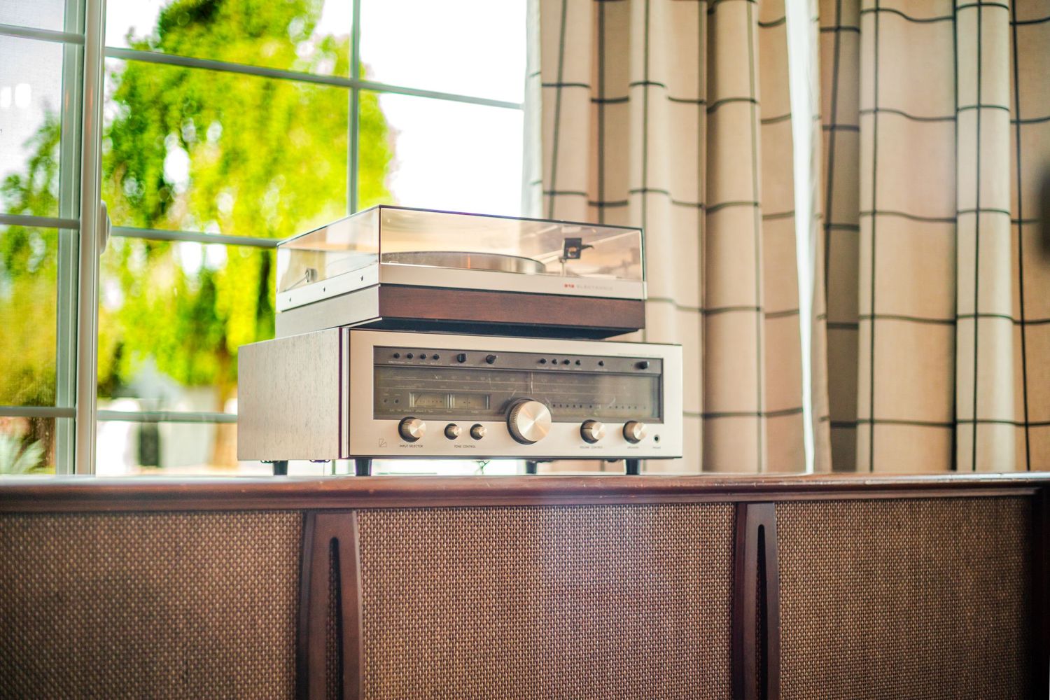 Vintage stereo receiver sits atop wooden cabinet near window with green foliage outside.