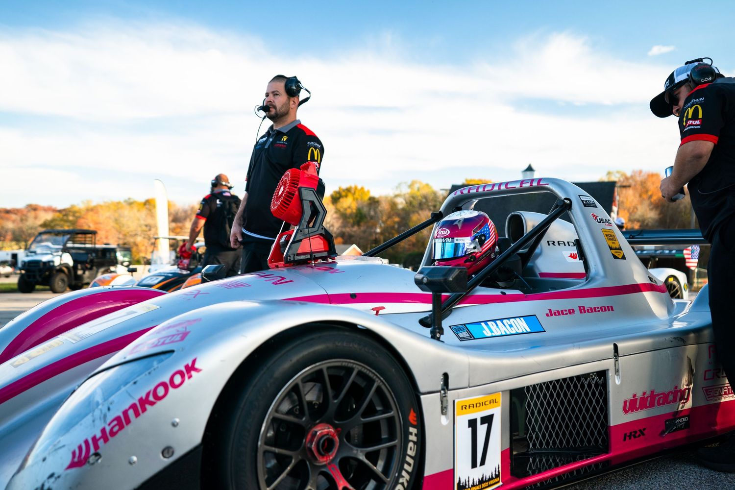 A race car driver wearing headphones and racing gear stands next to a pink and black formula-style race car on a track.