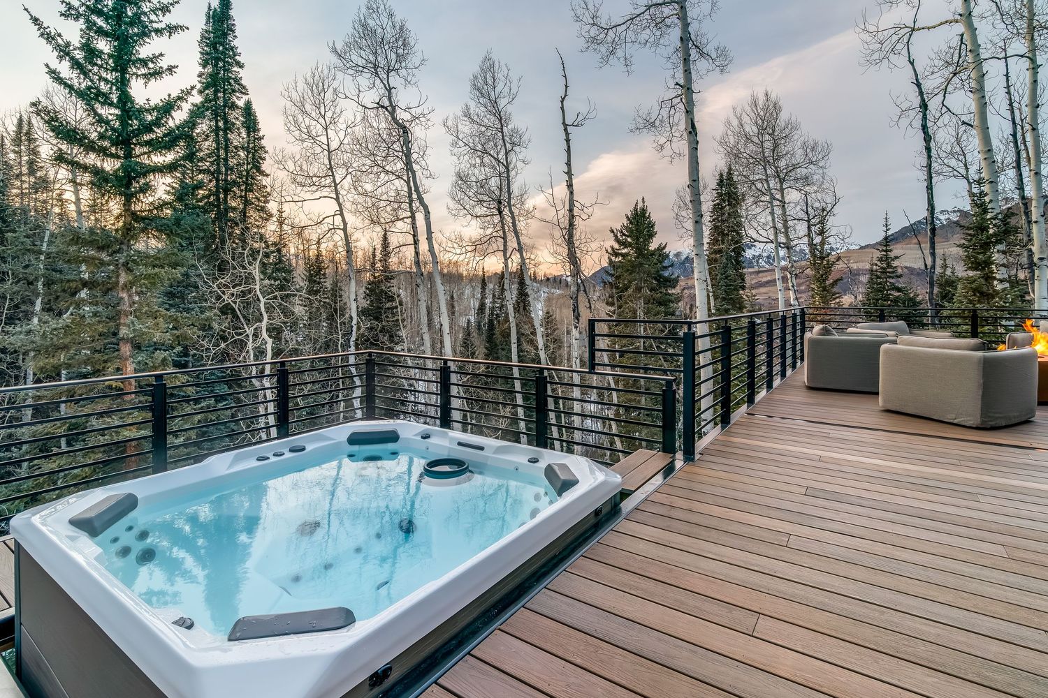 Hot tub on wooden deck with mountain views through cable railing at dusk.