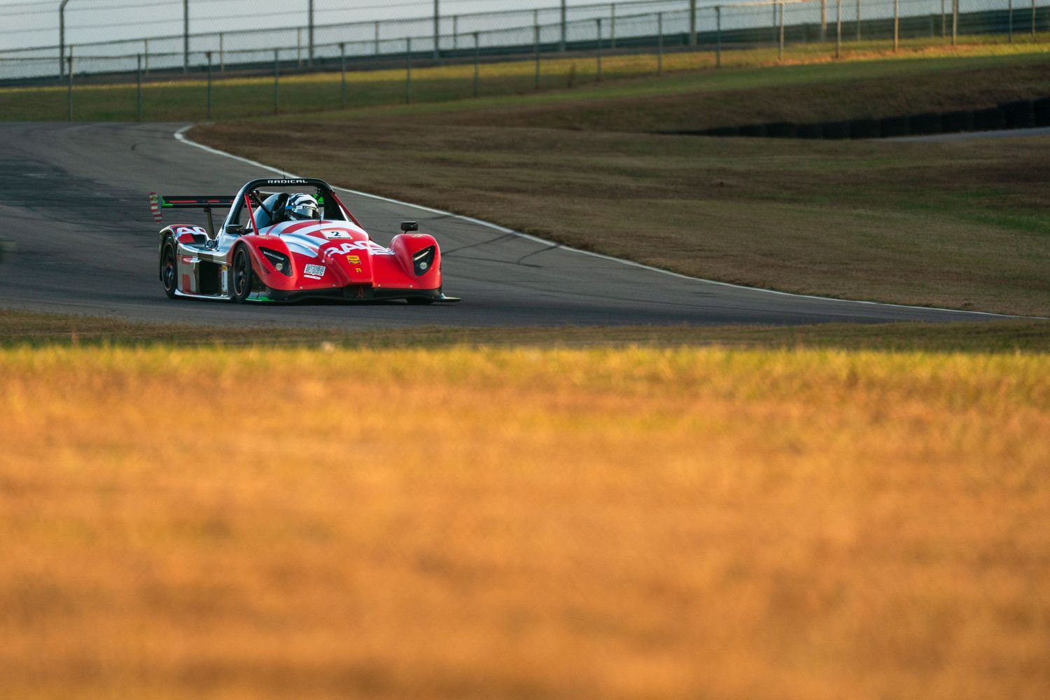 Racing car speeds past on track with golden sunlit grass in the foreground.
