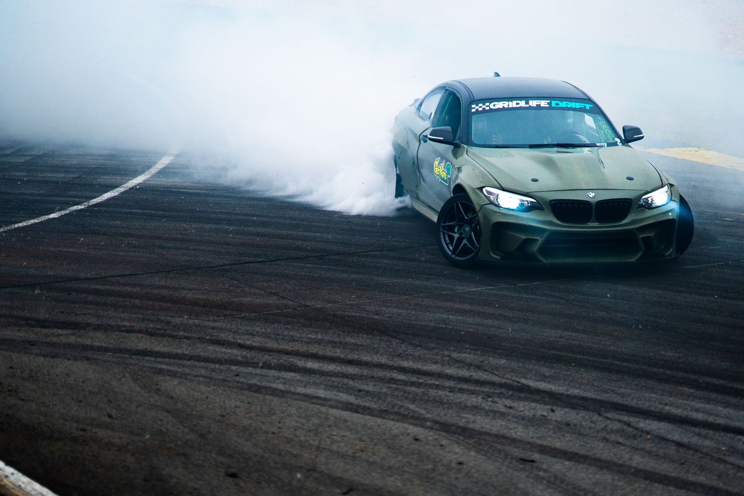 Olive green BMW M2 drifting with heavy tire smoke on a motorsport track during a drift event.