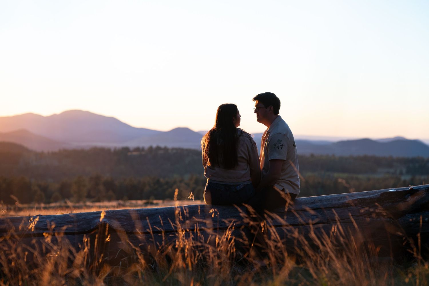 Couple sitting close together on a wooden fence at sunset, overlooking golden grass and distant mountains under a soft glowing sky.