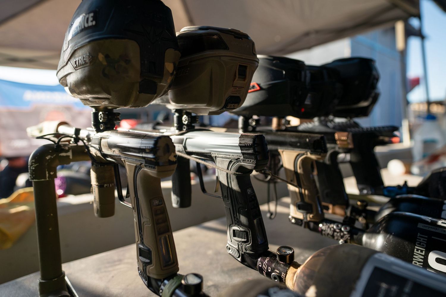 Row of paintball markers with hoppers and air tanks displayed on a rack at an outdoor paintball field.