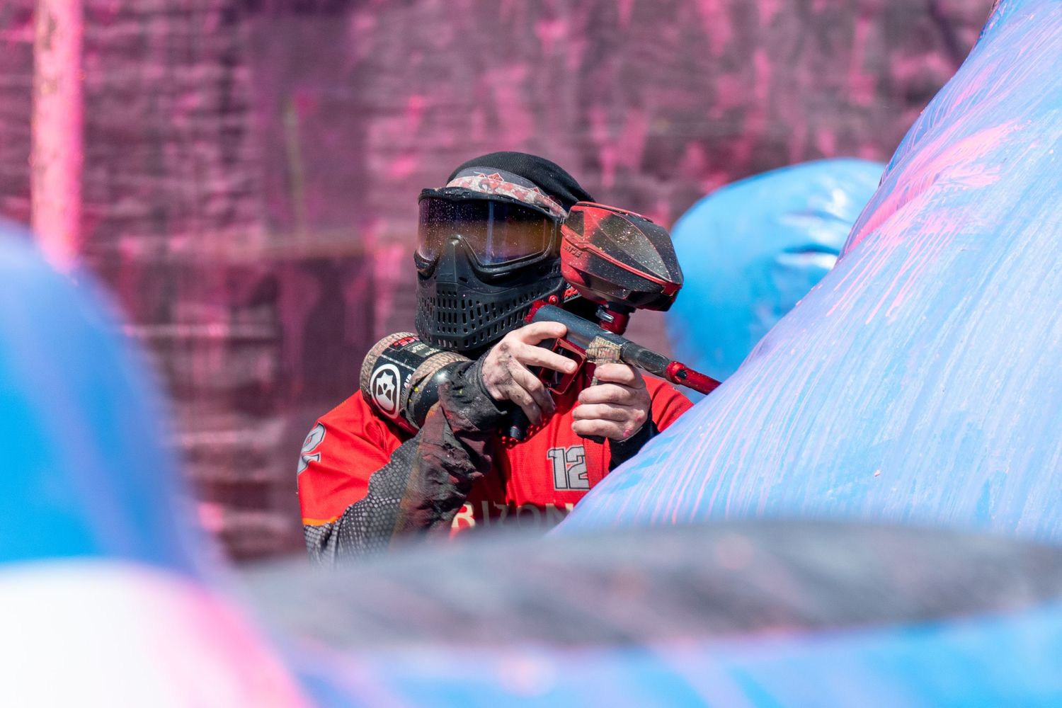 Two paintball players in red jerseys and protective masks crouch behind colorful inflatable bunkers during a match.