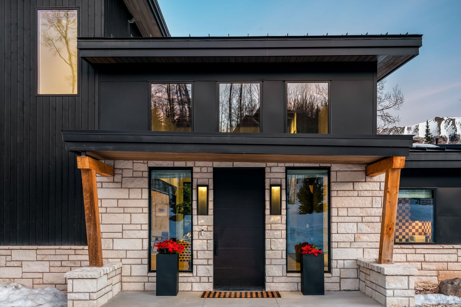 Modern home exterior with black siding, limestone accents, wood beams, and red potted plants flanking the front door.