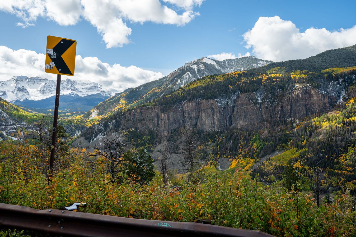 Yellow curve warning road sign with dramatic mountain cliffs and fall foliage in background.