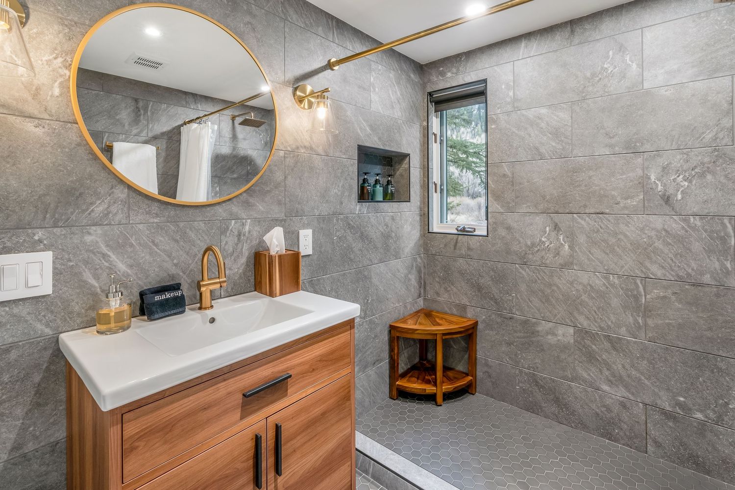 Modern bathroom with gray stone tiles, round gold mirror, wood vanity, and open walk-in shower with small wooden stool.