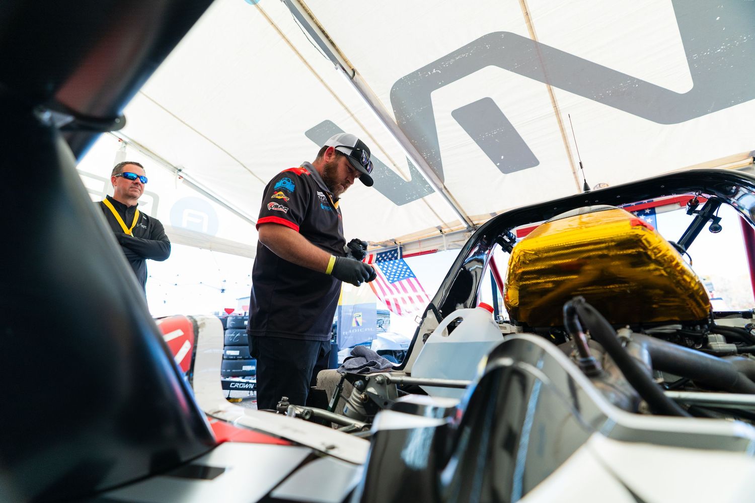 Racing crew members make final adjustments to vehicles under bright white canopy shelters.