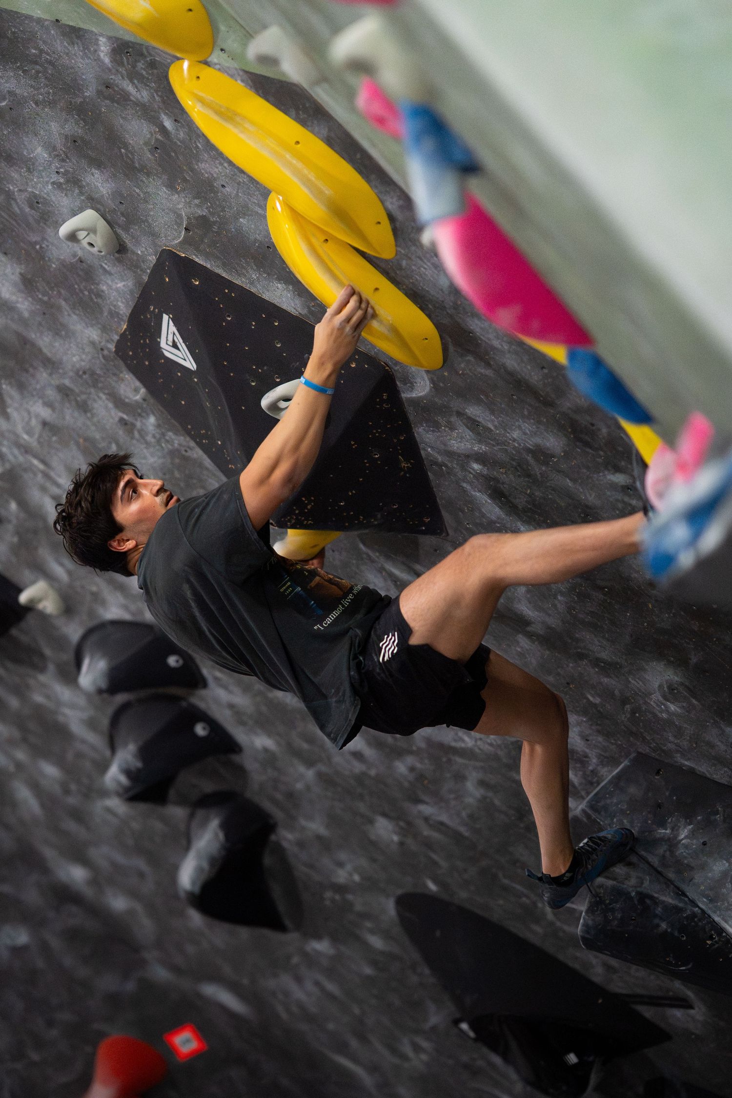 Climber in a gray shirt and black shorts reaches for a yellow hold while bouldering on a steep indoor gym wall.