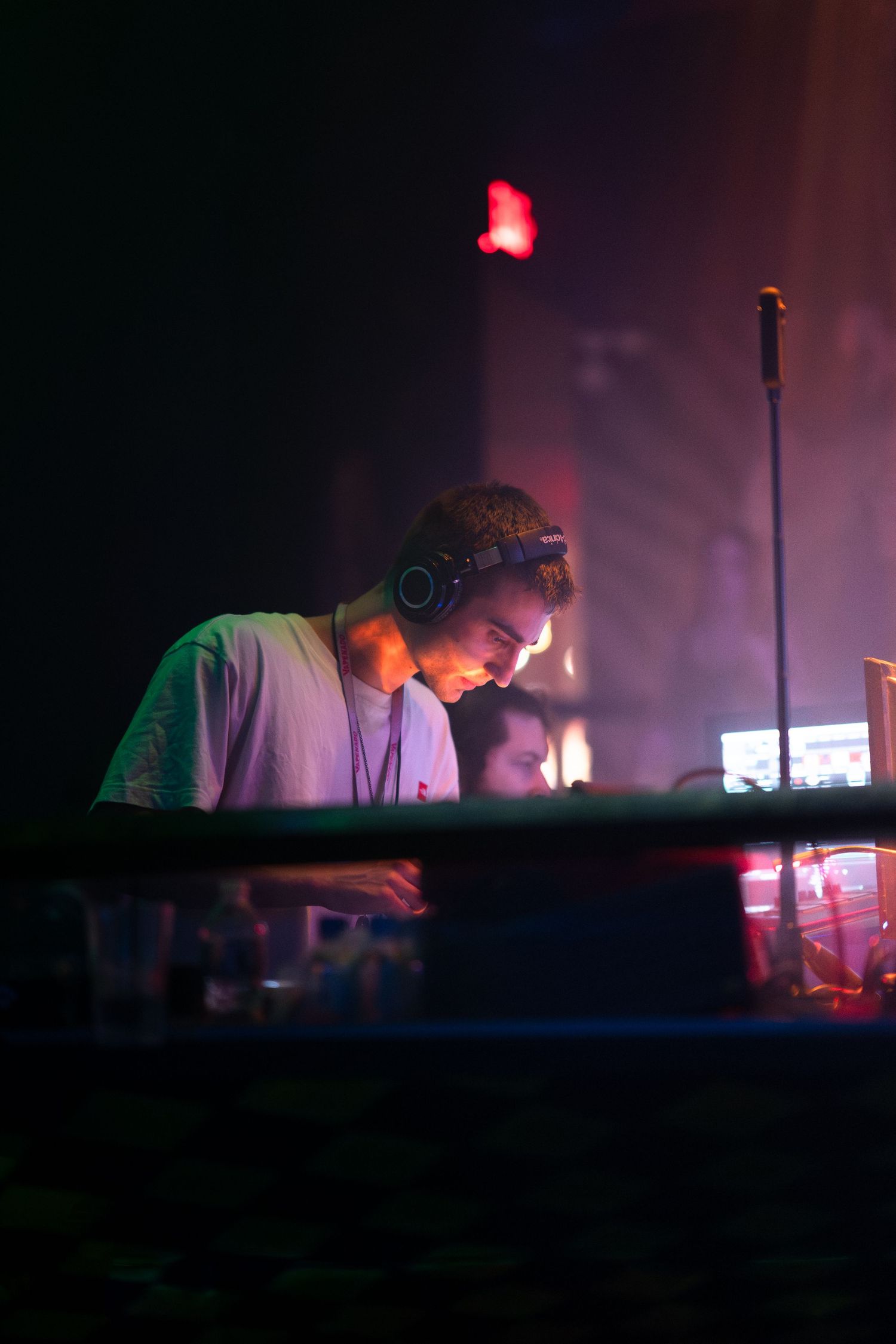 DJ performing at a nightclub wearing headphones while working at a dimly lit booth with red and purple lighting.