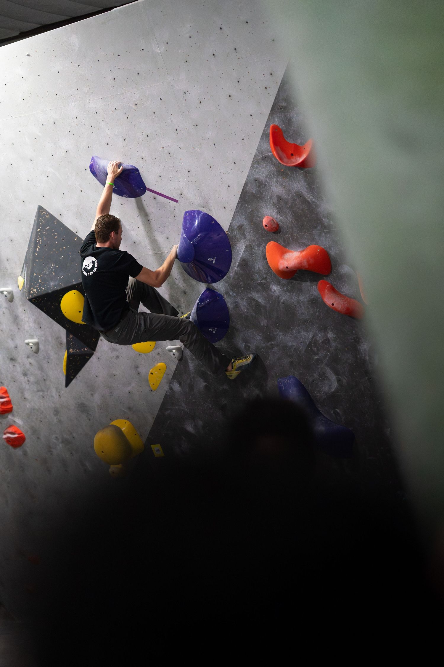 A climber in a black t-shirt reaches for a purple hold on an indoor bouldering wall with red and yellow holds.