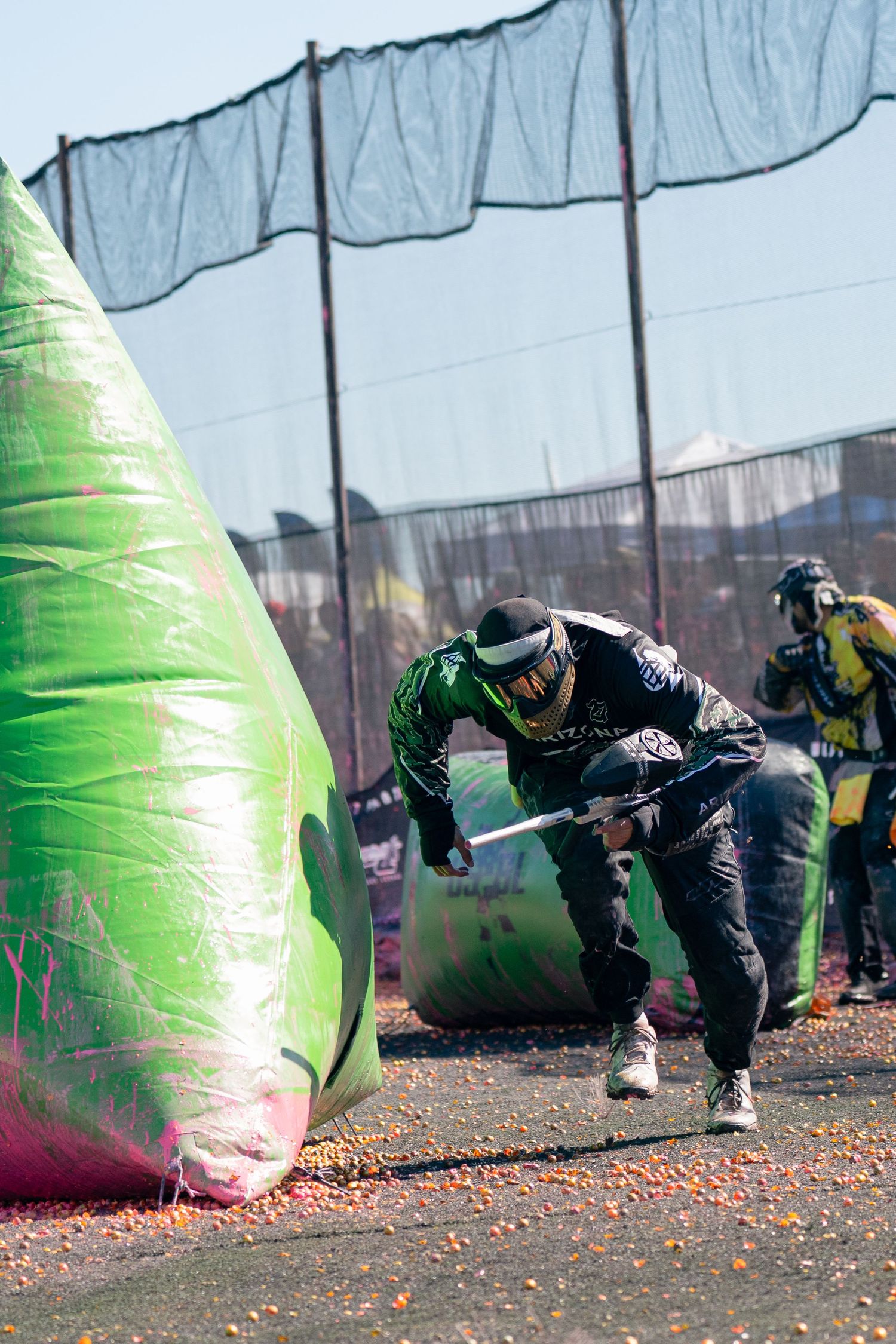 Paintball player in black gear crouching behind a green inflatable bunker during a competitive match.