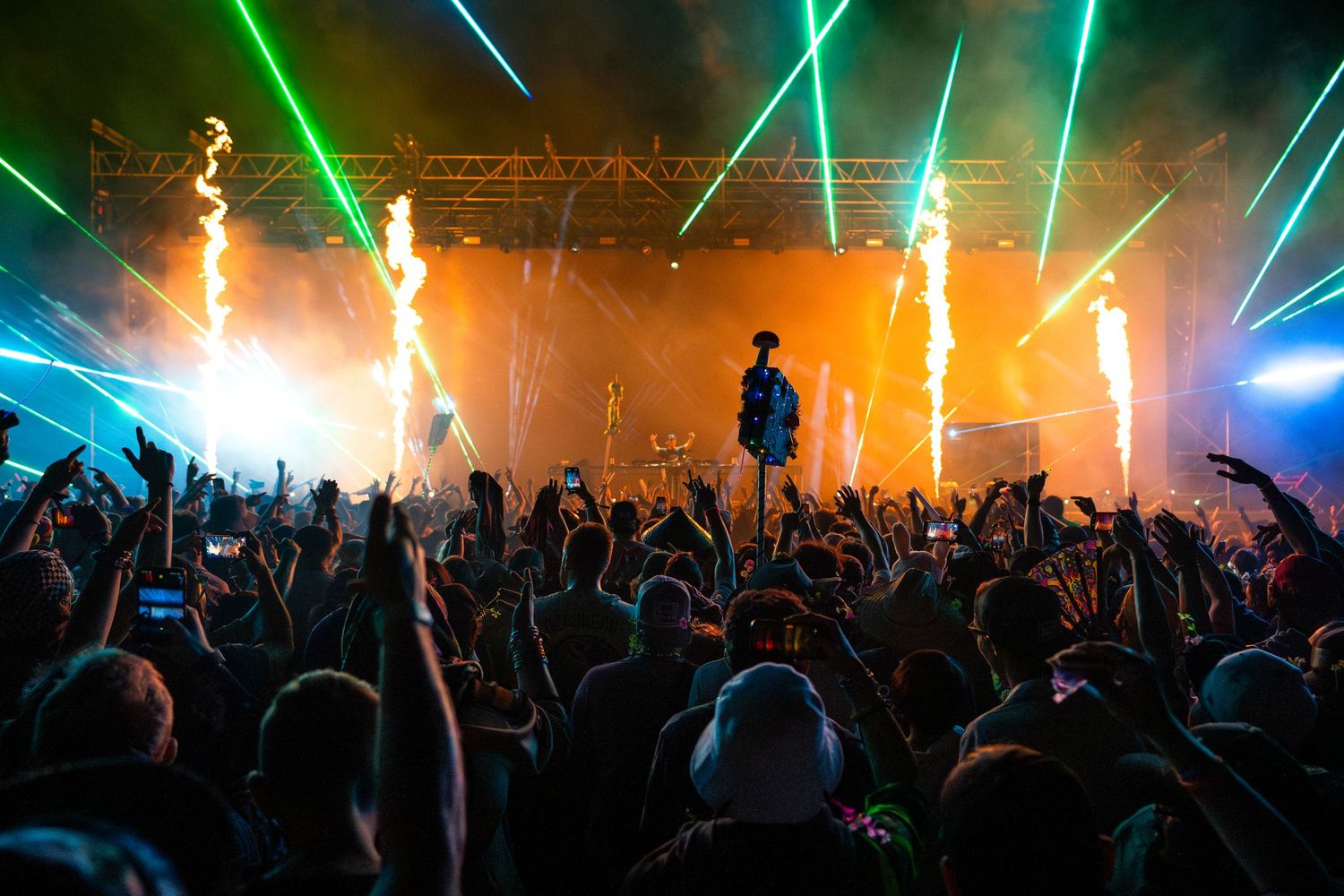 A performer on stage with pyrotechnics and green laser lights illuminating a massive crowd at an outdoor music festival.
