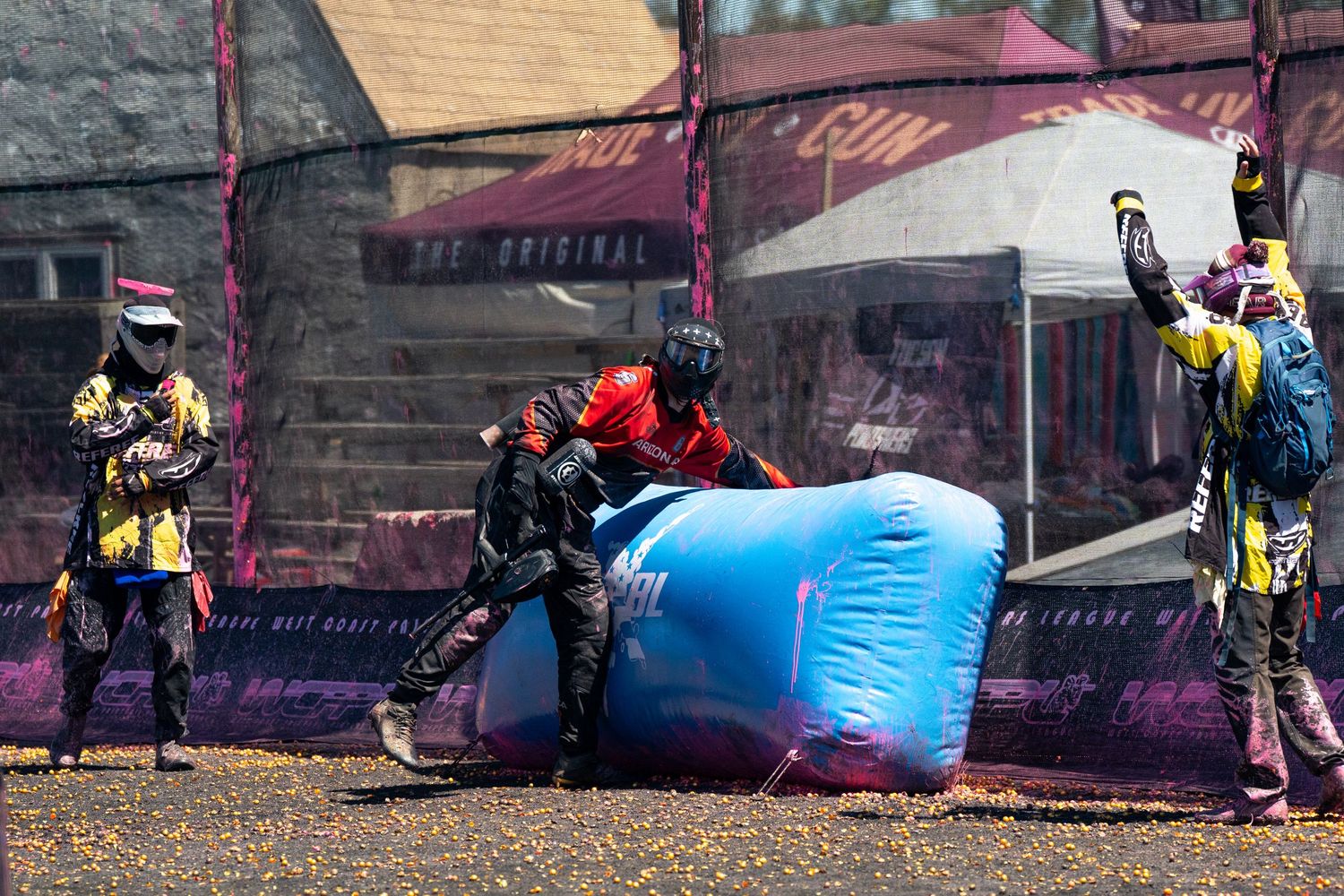 Players in protective gear compete in a paintball match, crouching behind a blue inflatable bunker on an outdoor field.