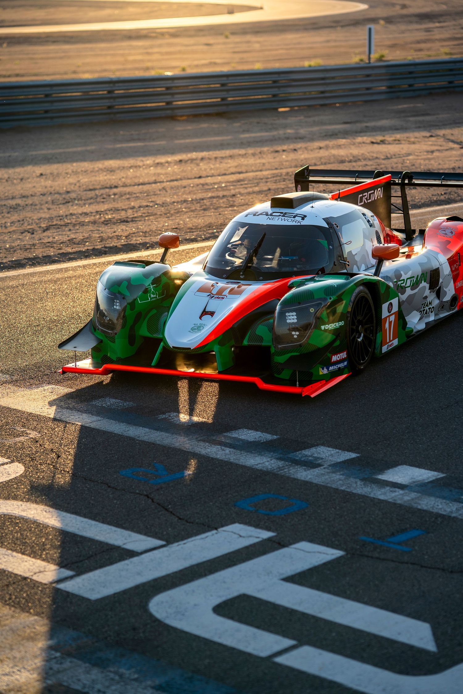 Racing prototype cars lined up at sunset on a race track with dramatic lighting and shadows.