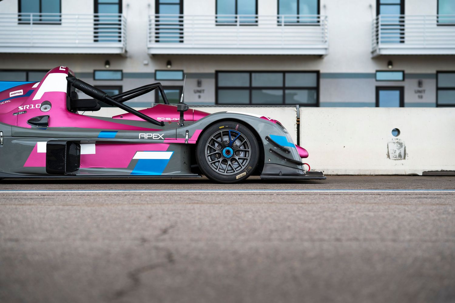 A pink and blue racing car speeds along a racetrack with modern buildings in the background.