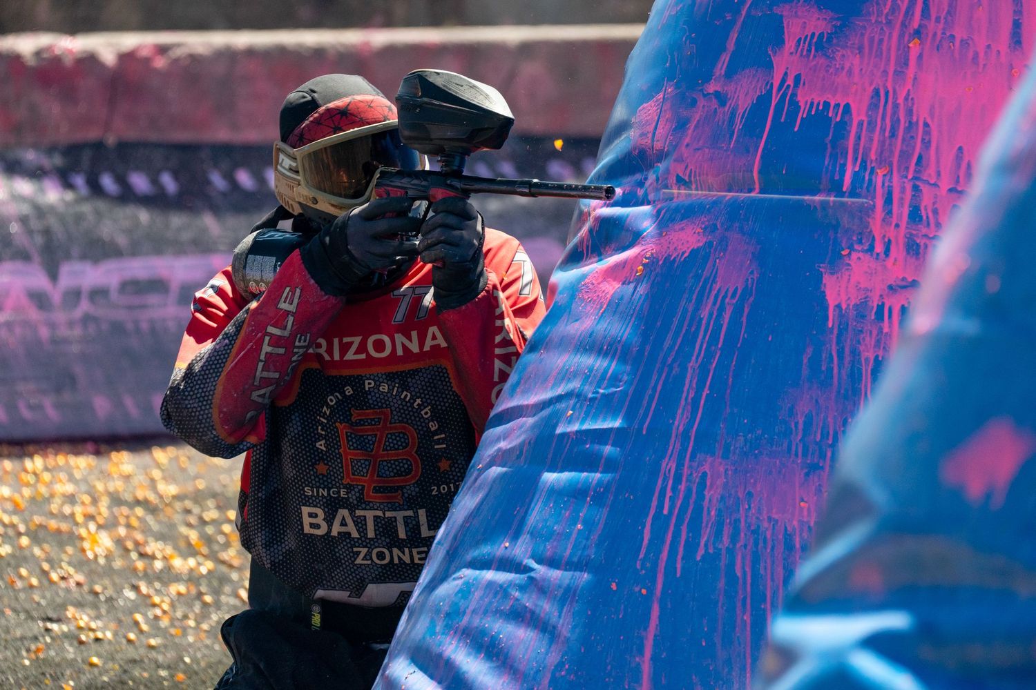 Paintball player in Arizona Battle Zone jersey aiming gun behind colorful splattered barrier during competitive match.