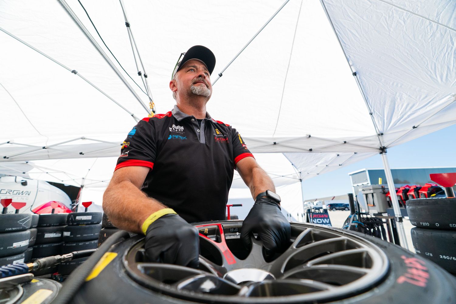 Mechanics perform maintenance work on race cars under white pop-up tents at a motorsport event.