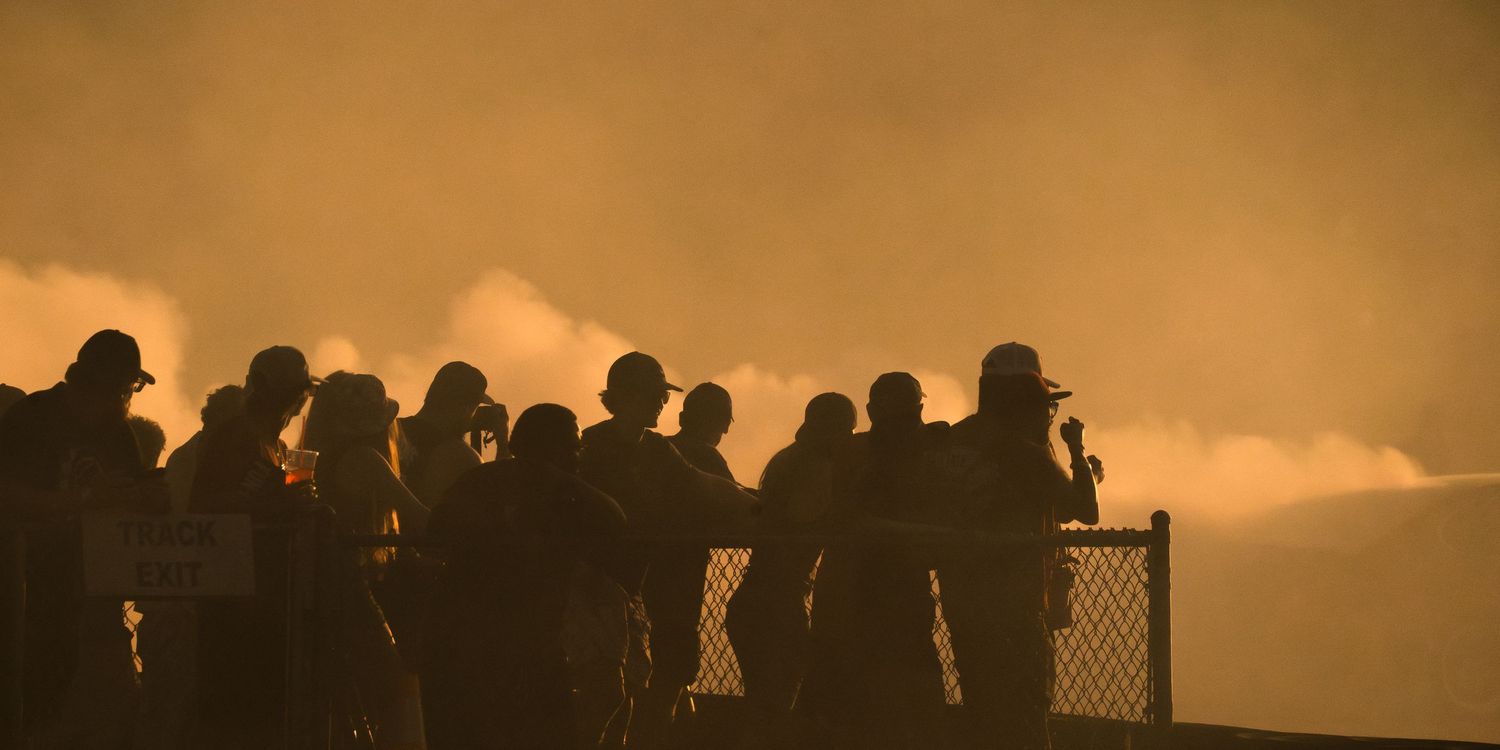 Silhouettes of spectators watching a dramatic smoky sunset at a racing event, backlit by golden orange skies.