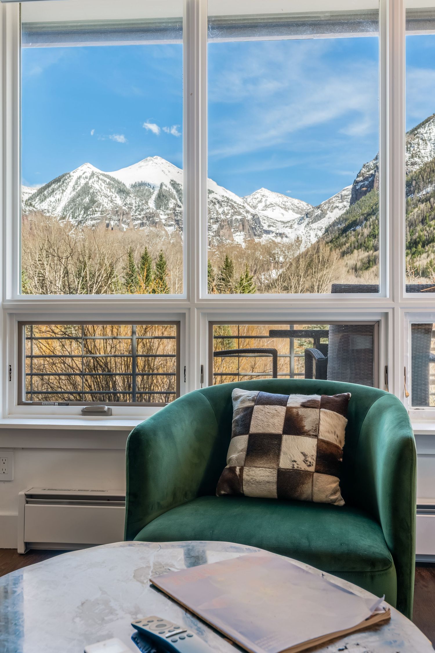 Green velvet armchair with checkered pillow beside a marble table, overlooking snow-capped mountains through large windows.