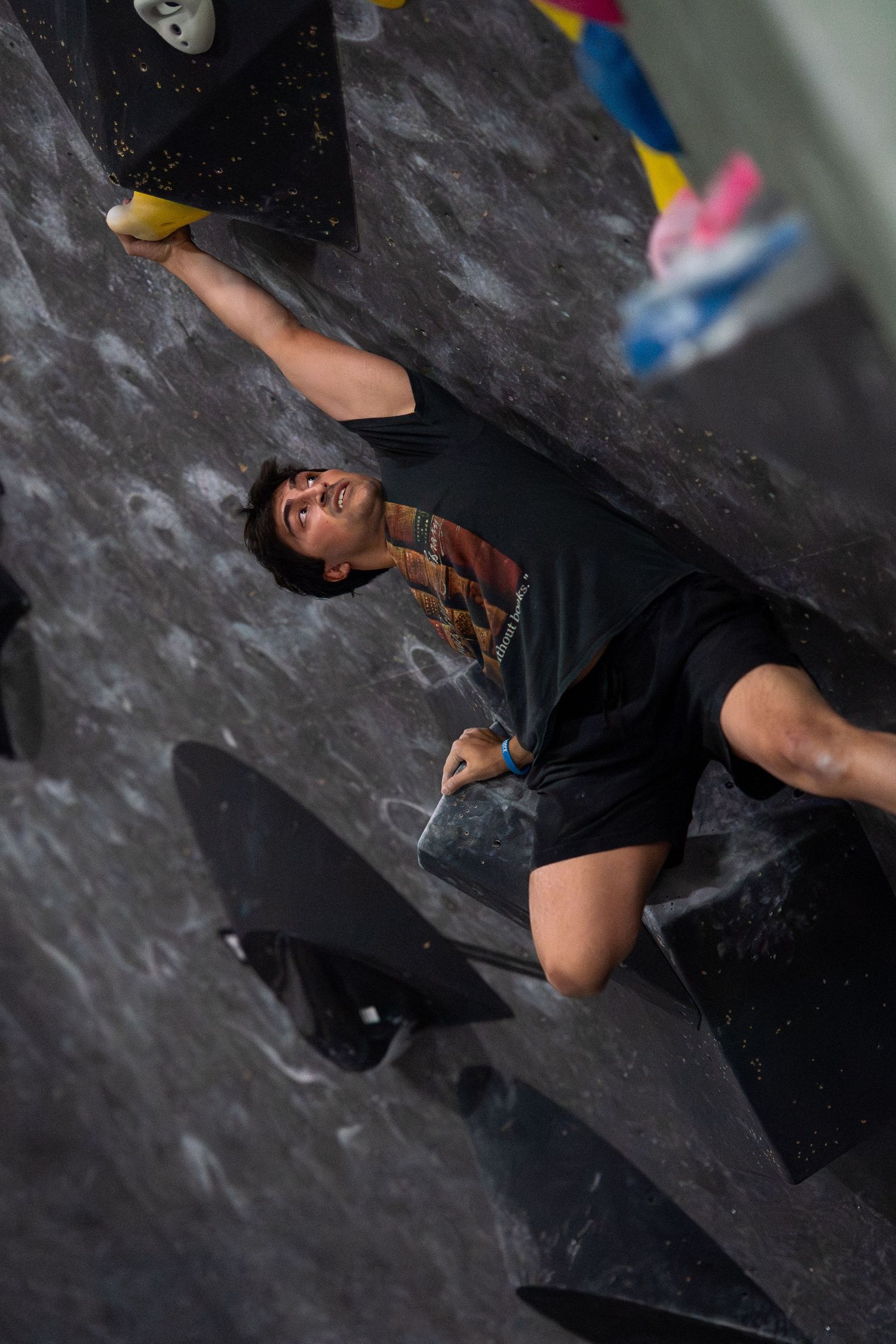 Climber in a black t-shirt and shorts reaches up with one arm while bouldering on a dark textured wall at an indoor climbing gym.