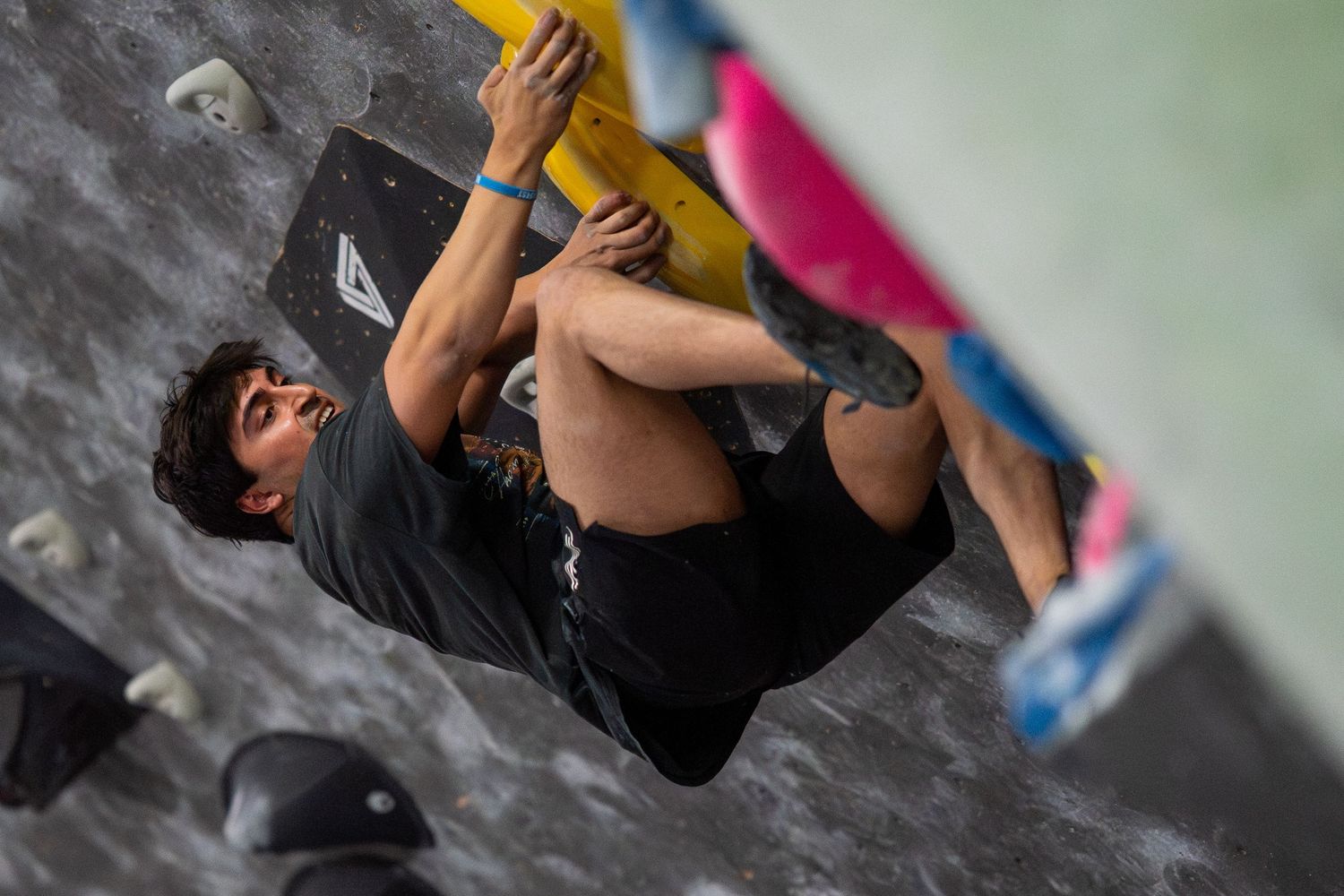 Climber in a black shirt and shorts hangs from a yellow hold while bouldering on an indoor gym wall with colorful holds.