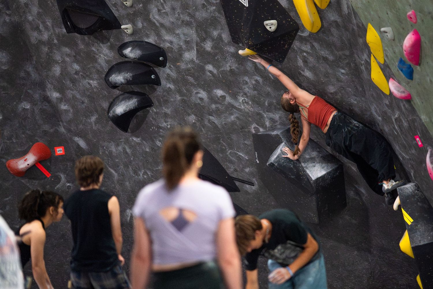 Climber in a red top stretches across a dark overhanging wall at an indoor bouldering gym while spectators watch from below.