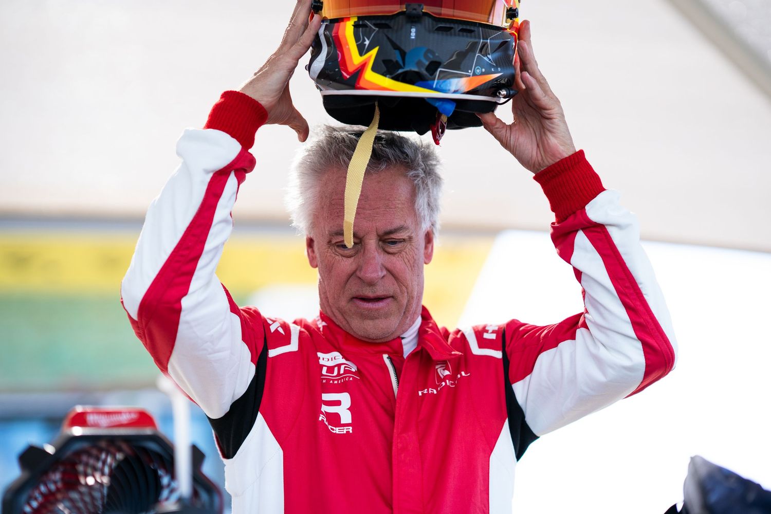 Race car driver in red suit adjusts helmet while preparing for competition at motorsport event.