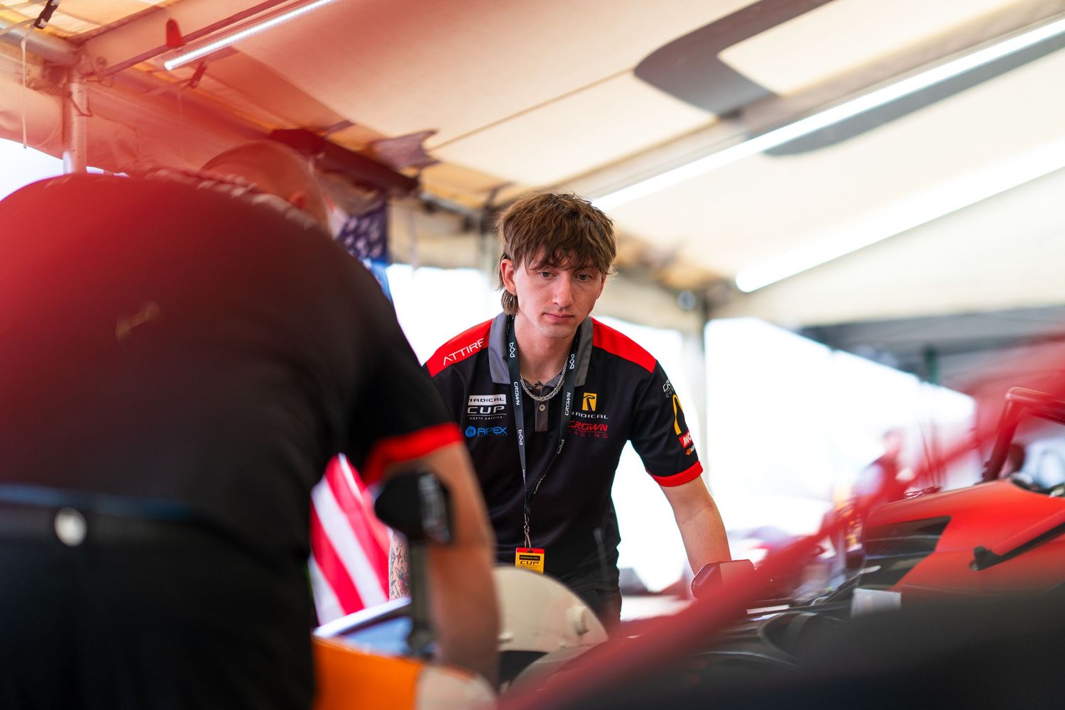 Race car driver preparing in the cockpit before a competition with American flag visible in background.